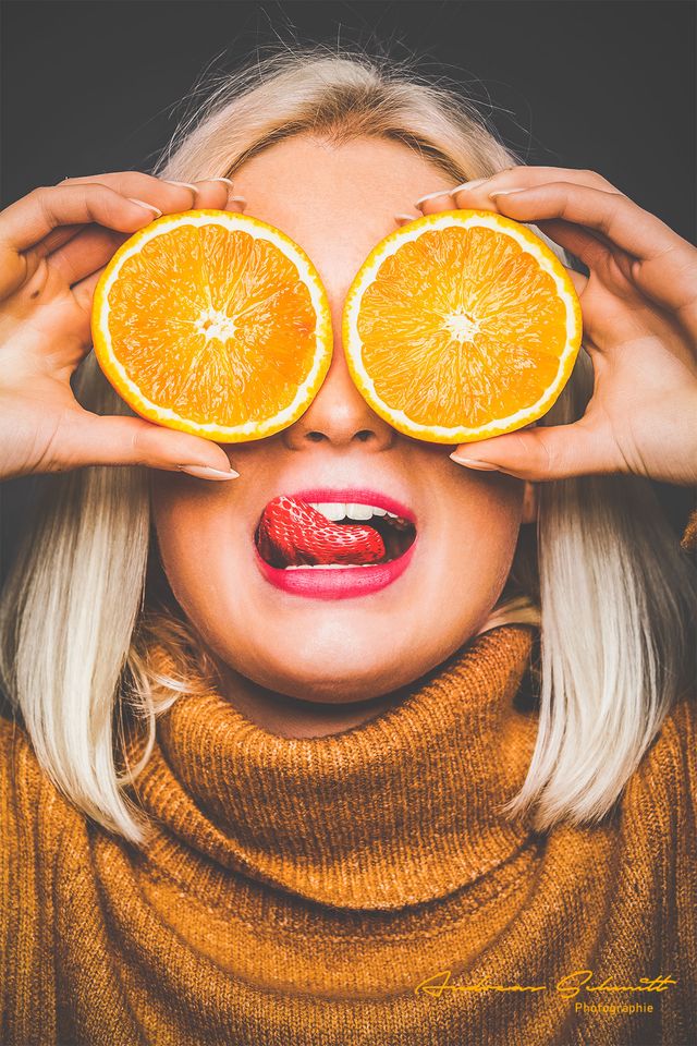 Blonde Frau hält Orangenscheiben vor die Augen und streckt die Zunge heraus